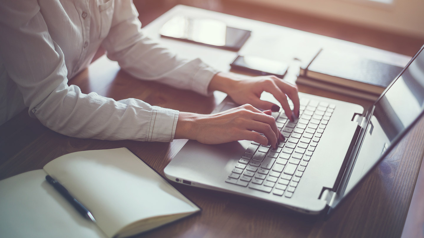 Woman wearing white shirt typing on laptop with book open to the side